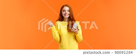 Happy girl share good news with you. Cheerful pretty redhead female holding smartphone, fist pump in triumph or celebration, winning in game, got message from guy she likes, orange background 98091284