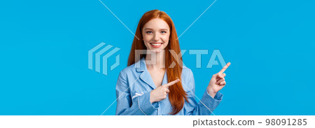 Satisfied cheerful redhead female in nightwear, pointing upper left corner, smiling friendly, help friend find place, perfect advertisement copy space, recommend product, standing blue background 98091285