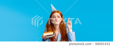 Eager and hopeful redhead european girl in nightwear, close eyes and cross fingers good luck, biting lip tempting or exciting, making wish hold birthday cake with lit candle, wear b-day cap 98091432