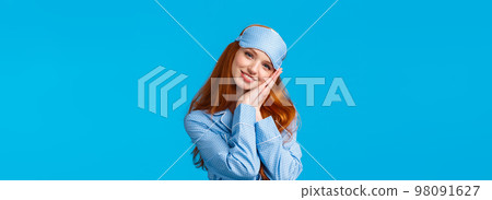 Have sweet dreams. Tender, lovely and feminine redhead teenage girl ready sleep tight, wearing sleeping mask and pyjama, lean on palms like pillow, smiling pleased and relaxed, blue background 98091627