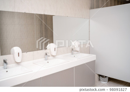 The interior of a public toilet. Row of wash basins with metal faucets on a marble slab, liquid soap dispensers, a long mirror on a gray wall The interior of a public toilet. Row of wash basins with metal faucets on a marble slab, liquid soap dispensers, a long mirror on a gray wall 98091827