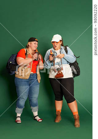 Portrait of two overweight women, friends in casual clothes with backpacks, eating hot dogs over green studio background Portrait of two overweight women, friends in casual clothes with backpacks, eating hot dogs over green studio background 98092220