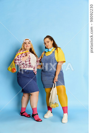 Portrait of two fat, beautiful women, friends posing with bag with fruits and vegetables over blue studio background. Diet 98092230