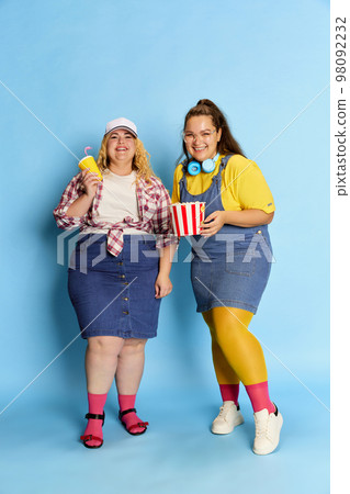 Portrait of two fat, beautiful women, friends posing with popcorn basket over blue studio background. Weekend leisure time 98092232