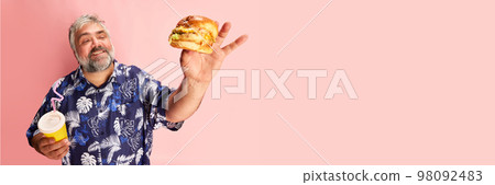 Portrait of mature man in colorful shirt smiling, posing with delicious burger over pink studio background. Fast food. Banner Portrait of mature man in colorful shirt smiling, posing with delicious burger over pink studio background. Fast food. Banner 98092483