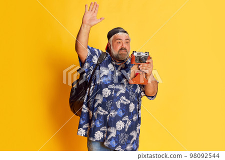 Portrait of fat mature man in stylish shirt posing, taking photo with vintage camera over bright yellow studio background 98092544