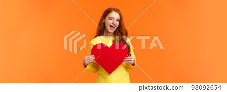 Hey gorgeous you have my heart. Valentines day, boyfriend and girlfriend concept. Cute and sassy redhead woman holding large sign and smiling, wink coquettish, standing orange background 98092654
