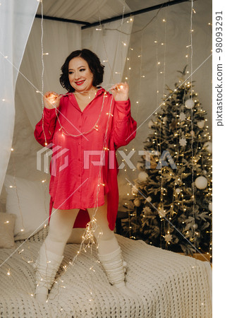 A woman in a red shirt is standing in the bedroom on a bed decorated for Christmas. Home New Year atmosphere A woman in a red shirt is standing in the bedroom on a bed decorated for Christmas. Home New Year atmosphere 98093291