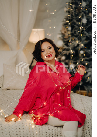 A woman in a red shirt in a room decorated for Christmas is sitting on a bed. Home New Year atmosphere A woman in a red shirt in a room decorated for Christmas is sitting on a bed. Home New Year atmosphere 98093338