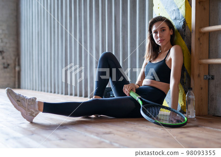 Portrait of female tennis player with racket sitting on gym floor 98093355