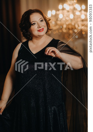 An elegant adult woman in a black cocktail dress poses in the interior. Fashionable portrait of a woman An elegant adult woman in a black cocktail dress poses in the interior. Fashionable portrait of a woman 98093383