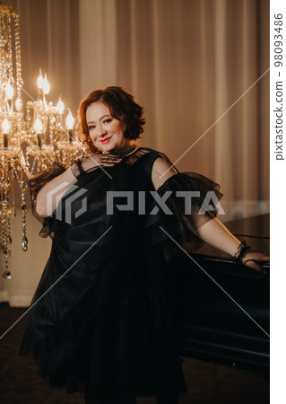 An elegant adult woman in a black cocktail dress poses in the interior. Fashionable portrait of a woman An elegant adult woman in a black cocktail dress poses in the interior. Fashionable portrait of a woman 98093486