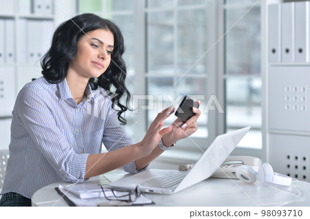 Beautiful young woman with phone working in office Beautiful young woman with phone working in office 98093710