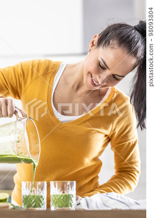 A woman pours the finished spinach smoothie into two glasses from a blender A woman pours the finished spinach smoothie into two glasses from a blender 98093804