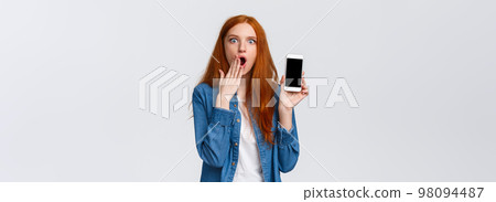 Impressed and astonished redhead woman seeing something unbelievable, spilling tea in internet, gasping cover opened mouth and showing samrtphone display, white background 98094487