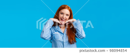 Lovely, charismatic redhead girl getting ready sleep, wearing nightwear, holding hands on jawline as showing her pure clean face, apply night cosmetics, skincare routine products, blue background 98094854
