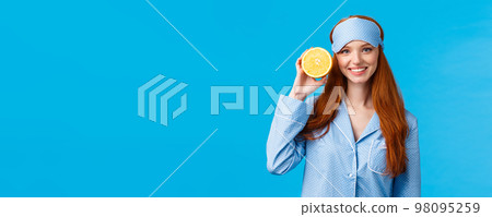 Start your morning right. Diet, healthy lifestyle and beauty concept. Cheerful glamour redhead woman in nightwear, holding slice orange near face, smiling, wearing sleep mask and pyjama Start your morning right. Diet, healthy lifestyle and beauty concept. Cheerful glamour redhead woman in nightwear, holding slice orange near face, smiling, wearing sleep mask and pyjama 98095259