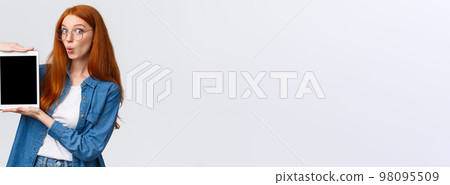 Amused, excited good-looking redhead woman in denim shirt, folding lips in wow sound, looking amazed camera, holding digital tablet, showing device screen, standing white background 98095509