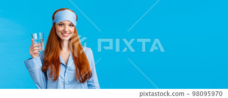 Waist-up shot glamour redhead woman taking care health, eating healthy, drinking water, wearing sleep mask and nightwear, holding glass and smiling energized, standing blue background 98095970
