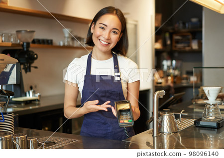 Portrait of asian barista girl at counter, showing card machine to client who wants to pay contactless, taking order, standing in cafe 98096140