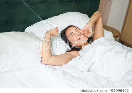 Happy mornings. Beautiful asian girl wakes up in a hotel suit, lying in white bed, stretching her arms and smiling happily Happy mornings. Beautiful asian girl wakes up in a hotel suit, lying in white bed, stretching her arms and smiling happily 98096192