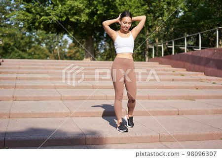 Sportswoman jogging outdoors. Smiling asian fitness girl in wireless earphones, listening music and running on road, workout 98096307