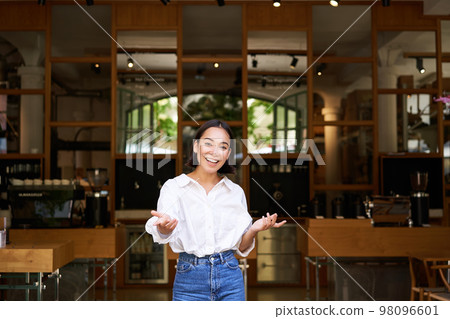Enthusiastic young businesswoman, asian girl showing her business, raising hands up and smiling, standing in front of restaurant Enthusiastic young businesswoman, asian girl showing her business, raising hands up and smiling, standing in front of restaurant 98096601