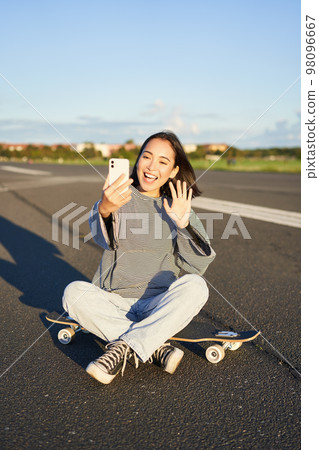 Happy teen asian girl takes selfie, video chats on smartphone app while sits on skateboard, skater records vlog while sits on longboard Happy teen asian girl takes selfie, video chats on smartphone app while sits on skateboard, skater records vlog while sits on longboard 98096667