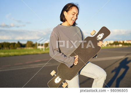 Beautiful asian teen girl playing with her longboard, holding skateboard as if playing guitar, standing on road on sunny day Beautiful asian teen girl playing with her longboard, holding skateboard as if playing guitar, standing on road on sunny day 98096805