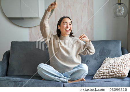 Happy and carefree girl singing and listening to music on smartphone app, using mobile phone as microphone, sitting on sofa 98096816