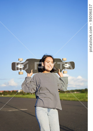 Vertical shot of carefree asian girl with longboard. Young woman skater holding cruiser on her shoulders and walking on road, skateboarding Vertical shot of carefree asian girl with longboard. Young woman skater holding cruiser on her shoulders and walking on road, skateboarding 98096837