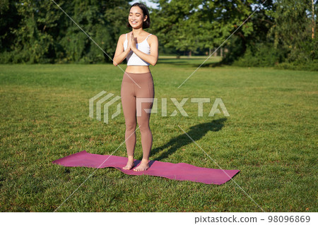 Mindfulness and wellbeing. Young woman doing yoga, standing on mat in park, making asana, meditating on fresh air, urban life concept 98096869