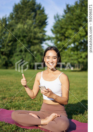 Vertical shot of asian woman shows thumbs up, recommending yoga training online, meditation app, doing exercises on fresh air in park Vertical shot of asian woman shows thumbs up, recommending yoga training online, meditation app, doing exercises on fresh air in park 98096871