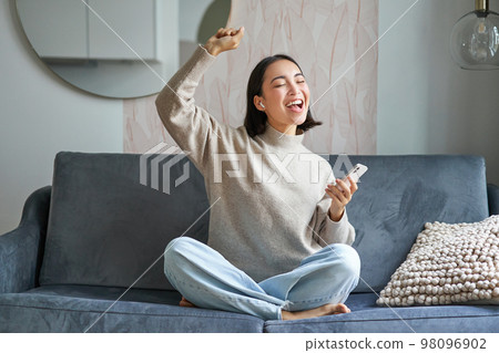 Portrait of carefree asian woman singing and listening music from smartphone app, using wireless headphones, smiling pleased, sitting on sofa at home 98096902