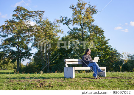 Portrait of young student, girl using laptop, sitting in park on bench, typing on computer 98097092