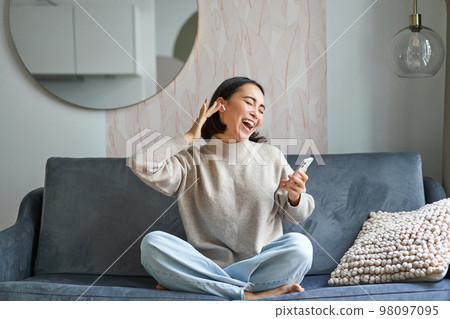 Portrait of carefree asian woman singing and listening music from smartphone app, using wireless headphones, smiling pleased, sitting on sofa at home Portrait of carefree asian woman singing and listening music from smartphone app, using wireless headphones, smiling pleased, sitting on sofa at home 98097095