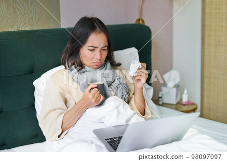 Portrait of asian girl sneezing and wiping runny nose, feeling sick, staying in bed with influenza, looking at laptop 98097097