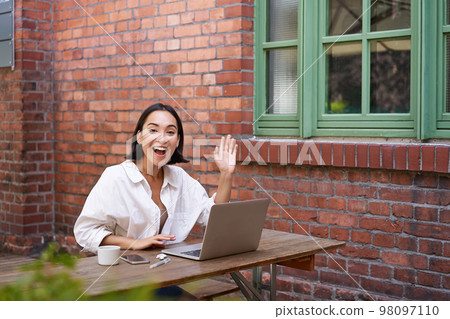 Friendly asian woman sitting with laptop, waving at you, saying hello, hi gesture, greeting you while working with computer 98097110