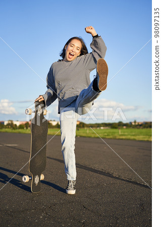 Excited asian girl dancing, standing with skateboard, skating on longboard and shouting carefree Excited asian girl dancing, standing with skateboard, skating on longboard and shouting carefree 98097135