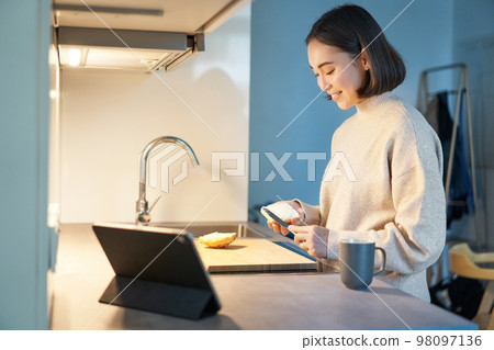Portrait of young korean woman, making toast and watching video on digital tablet, looking at screen Portrait of young korean woman, making toast and watching video on digital tablet, looking at screen 98097136