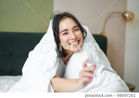 Beautiful asian woman sitting on bed, covered with white duvet, smiling, enjoying happy weekend morning, laughing at camera 98097202