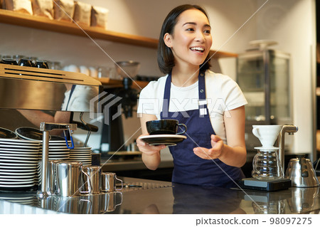 Portrait of beautiful asian girl, student working part-time in cafe, holding cup of coffee, made order, looking for client, wearing uniform Portrait of beautiful asian girl, student working part-time in cafe, holding cup of coffee, made order, looking for client, wearing uniform 98097275