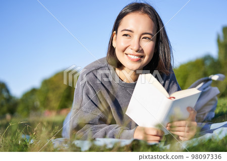 Portrait of cute korean girl, reading in park while lying on grass, relaxing with favorite book in hands, smiling happily Portrait of cute korean girl, reading in park while lying on grass, relaxing with favorite book in hands, smiling happily 98097336