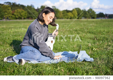 Smiling asian girl learns how to play ukulele via laptop, online video tutorials, sitting on grass in park with musical instrument 98097362