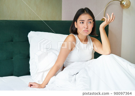 Portrait of puzzled asian woman lying in bed covered with warm duvet blanket, looking confused, shrugging with clueless face 98097364