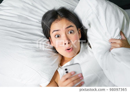 Portrait of asian girl wakes up in morning, looks at mobile phone with shocked face, overslept, hugging pillow and grimacing 98097365
