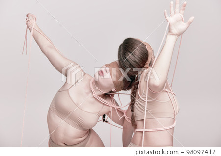 Connection. Portrait of two young girls posing in underwear and ropes with eyes closed over light studio background. Concept of weird people, contemporary photography 98097412