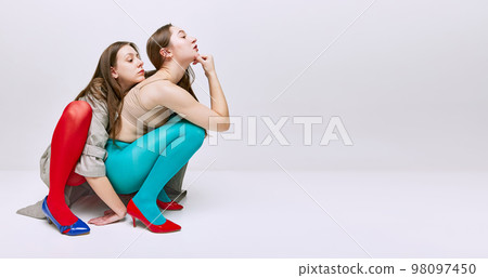 Portrait of two young girls in bright red and blue tights and coat posing over grey studio background. Vivid style, queer, art, fashion. Banner, flyer 98097450