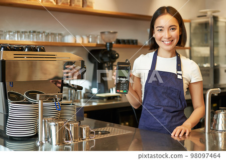 Smiling asian girl barista, cafe owner in apron, showing card machine, payment reader, taking contactless orders in her coffee shop Smiling asian girl barista, cafe owner in apron, showing card machine, payment reader, taking contactless orders in her coffee shop 98097464