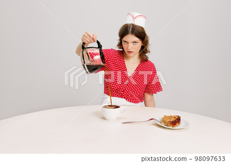 Stylish young female waitress in retro american fashion style of 70s, 80s sitting at table and drinking coffee over grey background. Funny meme emotions, ad, sales Stylish young female waitress in retro american fashion style of 70s, 80s sitting at table and drinking coffee over grey background. Funny meme emotions, ad, sales 98097633